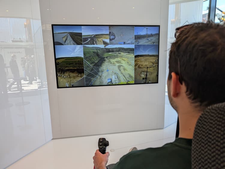 Mann sitzt vor einem Bildschirm auf dem Aufnahmen einer Baustelle zu sehen sind und steuert so einen Turmdrehkran